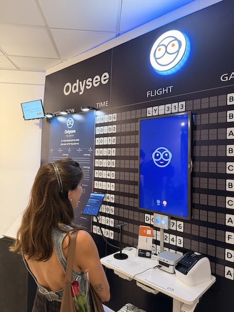 HE: מבקרת עומדת מול מיצב Odysee, גבה למצלמה, ומתבוננת במסך השיחה שמציג את פני Tico על רקע כחול. מעליה קיר לוח היציאות עם הכיתוב Odysee, סימן Tico מואר, וכותרות העמודות TIME ו-FLIGHT מעל אריחים מתהפכים. לפניה עמדת הדיבור הלבנה ועליה מיקרופון, מחשב ומדפסת מדבקות.