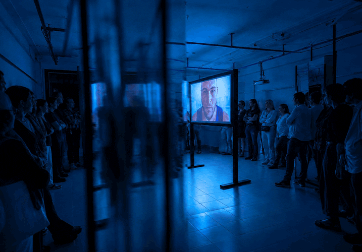 Visitors standing on both sides of a dark exhibition space, watching the video installation under blue projection lighting.